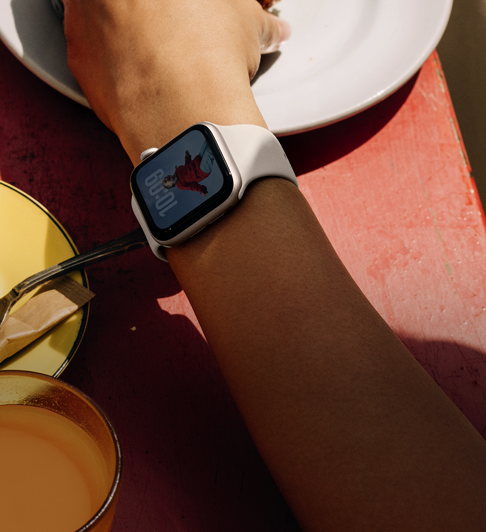 A person's arm grabbing a breakfast food, Apple Watch SE 3 on wrist, Starlight aluminum case, always-on display, portrait of a person on watch face, starlight Sport Band